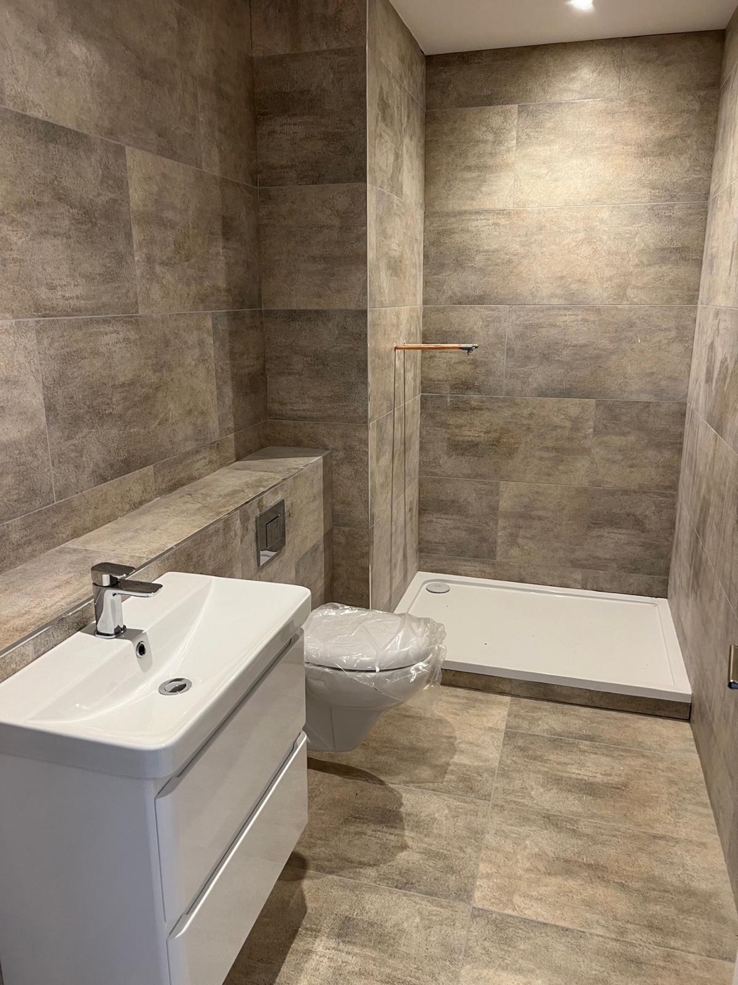 A stone-tiled shower room with a wall-hung basin unit and a rimless WC — the sort of refurbishment where the pipework has to be planned before a single tile is set out.
