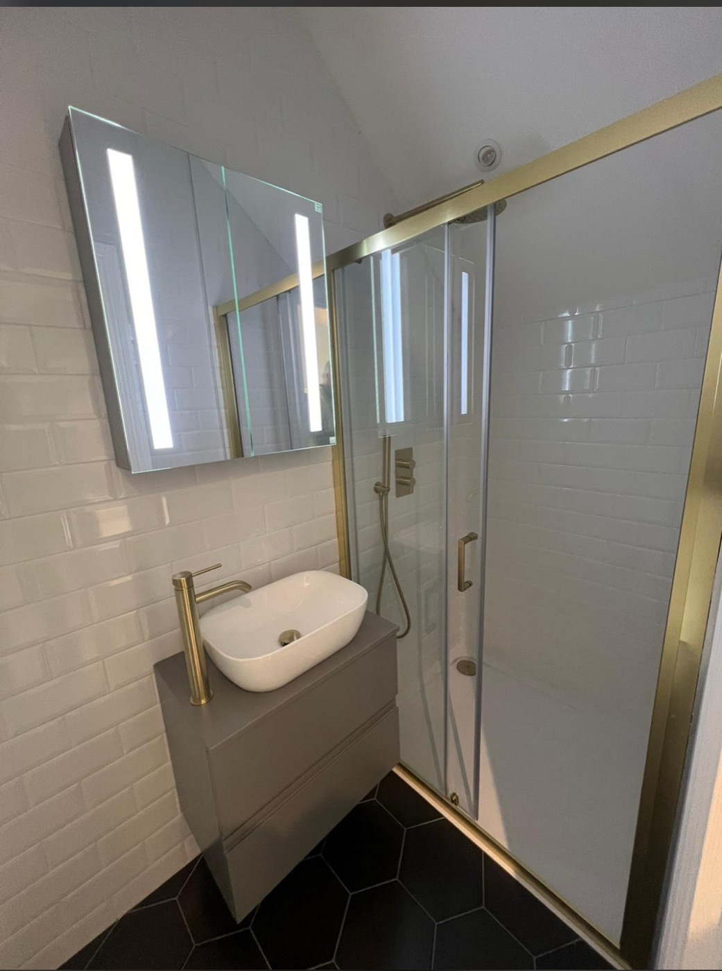 A brass-framed sliding shower enclosure, counter-top basin on a grey vanity and hexagonal floor tiles — a compact room planned so every centimetre earns its place.