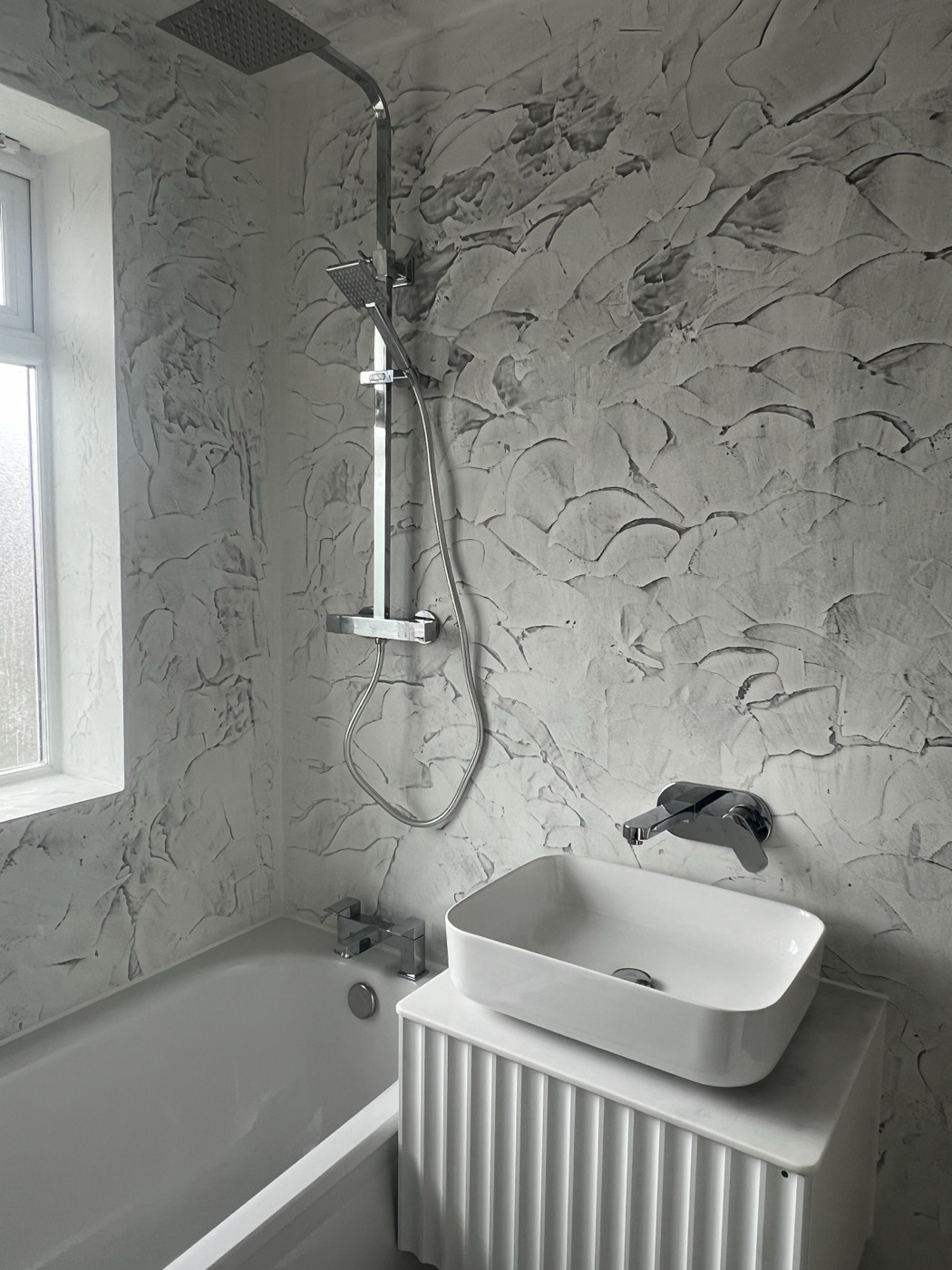 A bath with a thermostatic shower over it, textured wall finish and a basin mounted above the radiator — bathroom plumbing fitted around an awkward existing layout.